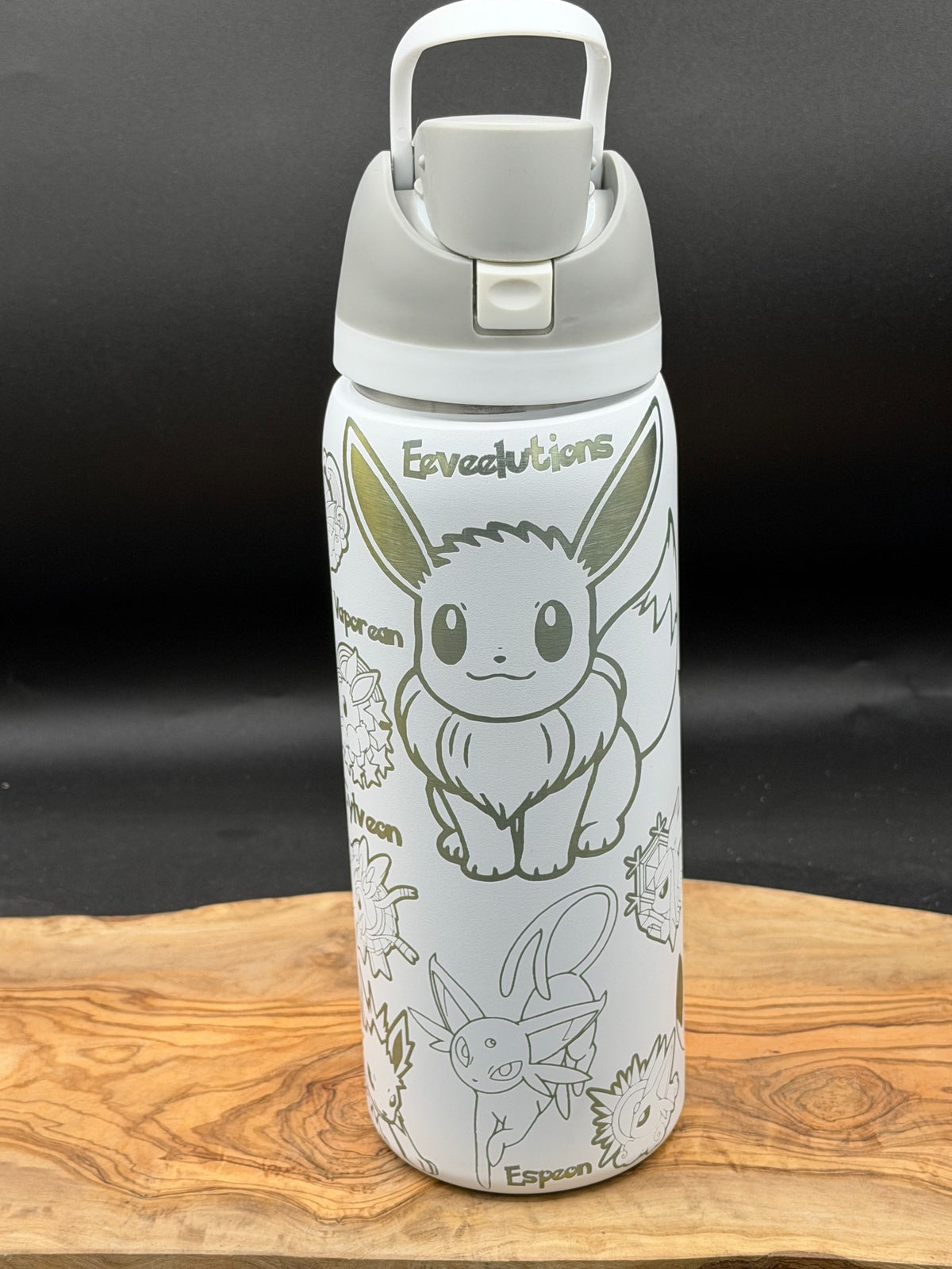 A white insulated water bottle with a gray lid and handle, decorated with Pokémon Eevee line art and text, displayed on a wooden surface against a dark background.
