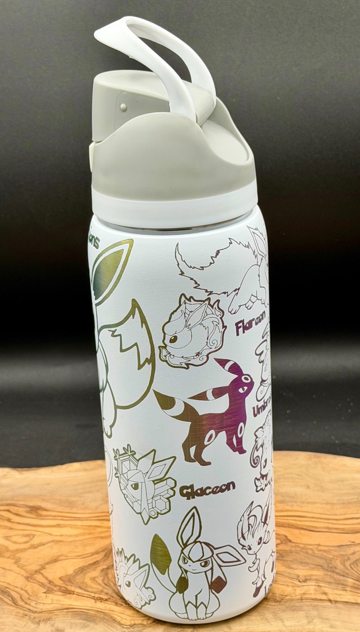 A white insulated water bottle with a gray lid and handle, decorated with various Eevee evolutionary line art illustrations in green, purple, and black, resting on a wooden surface against a dark background.