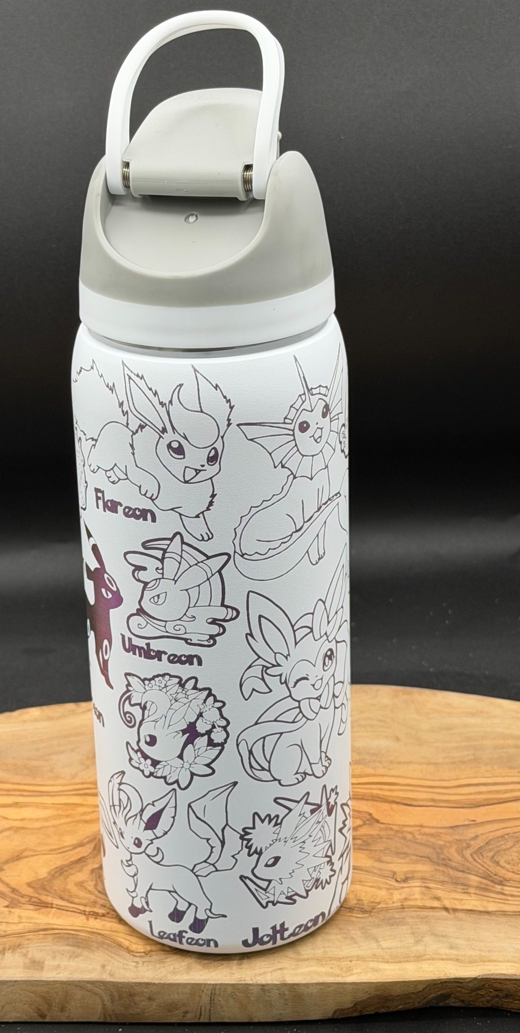 A white insulated water bottle with a gray lid and handle, decorated with black line art illustrations of various Eevee evolutions and names, displayed on a wooden board against a dark background.