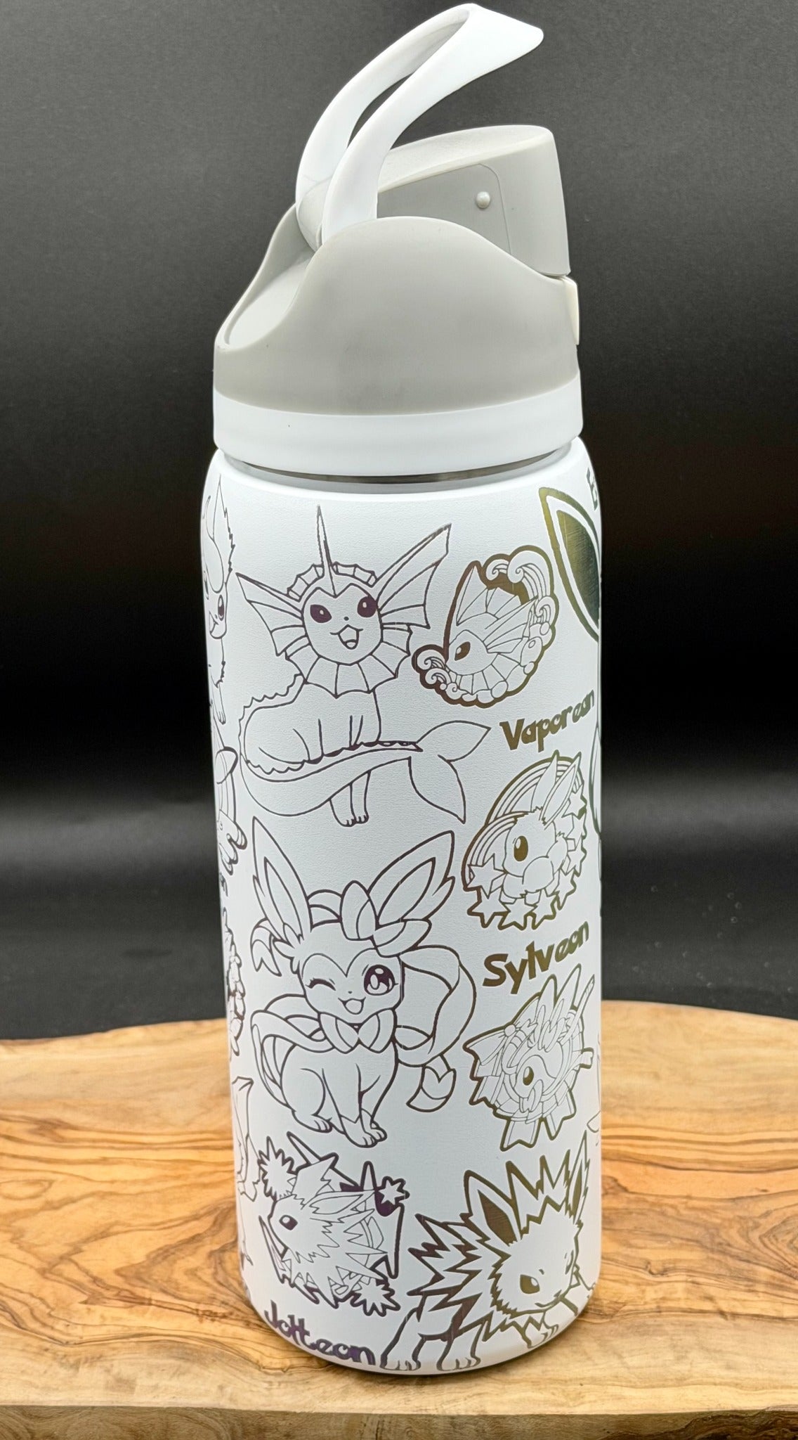A white insulated water bottle with a gray flip-top lid, decorated with black line art illustrations of various Pokémon characters and their names, resting on a wooden surface against a dark background.