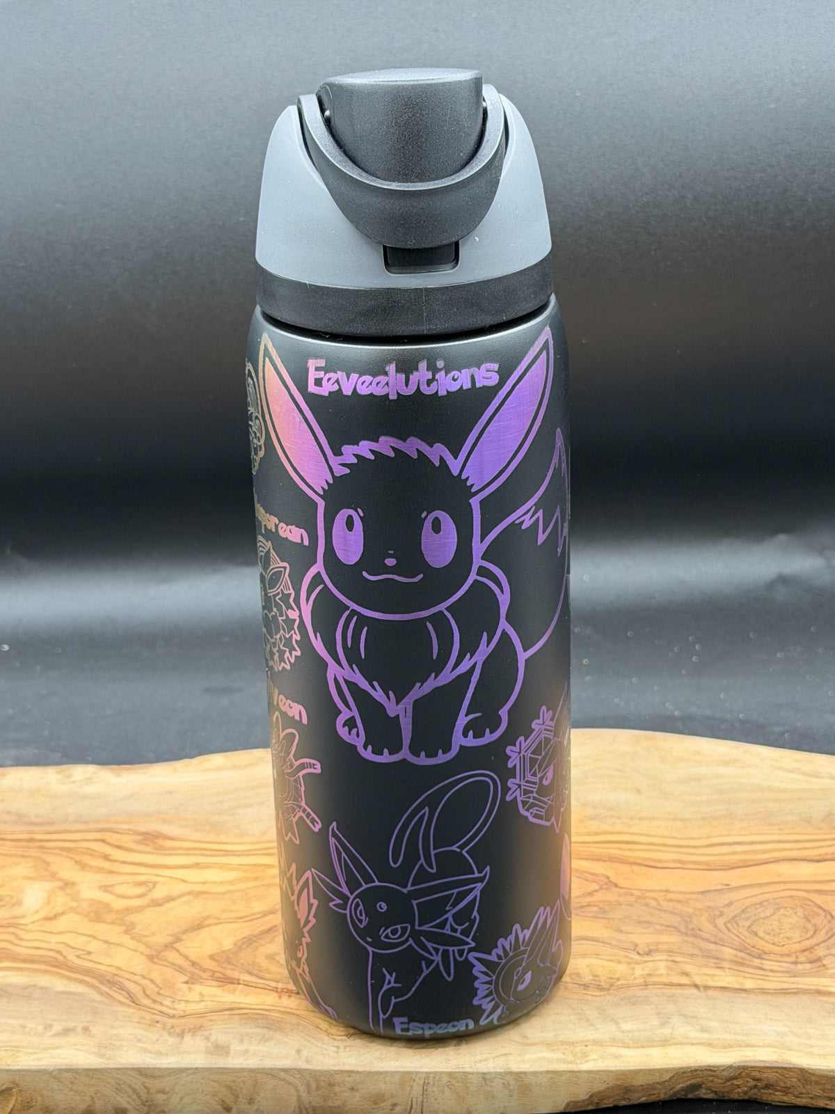 A black insulated water bottle with a gray flip-top lid, featuring purple line art illustrations of Eevee and the word 'Eeveevisions' on the front, set on a wooden surface against a dark gray background.